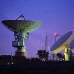 picture of 3 large satellites pointing up at space. it's nighttime and the sky looks purple. it's spotted with stars