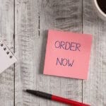 Handwriting text Order Now. Concept meaning the activity of asking for goods or services from a company Stationary placed next to a cup of black coffee above the wooden table.